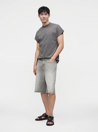 Jeune homme portant un débardeur gris, un short en jean gris clair et des sandales noires, debout avec une main dans la poche.