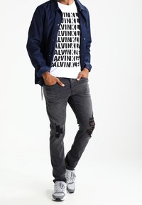 Mörkblå lättviktsjacka över en vit t-shirt med fet svart "CALVIN" tryck, kombinerad med slitna mörkgrå jeans och grå sneakers.