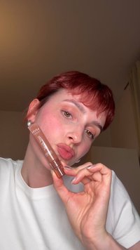 Person with short red hair holding a NYX professional makeup lip product near their face, wearing a white shirt indoors.