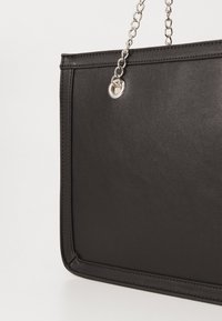 Geantă tote din piele sintetică neagră, cu o textură netedă, formă rectangulară și mânere din lanț argintiu, având un detaliu hardware circular.