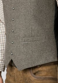 Graue Wollweste mit strukturierter Oberfläche, ausgestattet mit fünf metallischen Knöpfen und einer Seitentasche. Kombiniert mit einem braunen Ledergürtel und strukturierten braunen Hosen.