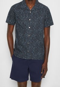 Koszulka z krótkim rękawem w ciemnoniebieskim materiale z jasnoniebieskim abstrakcyjnym wzorem, z zapięciem na guziki z przodu i kołnierzykiem w kształcie ostrego kąta.