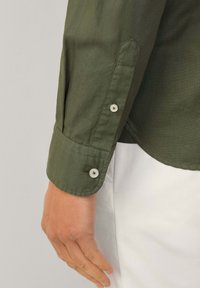 Camicia verde oliva a maniche lunghe con polsini con bottoni, texture morbida e leggera lucentezza. Presenta bottoni bianchi e una vestibilità sartoriale.