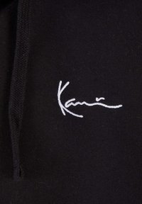 Black fabric with a smooth texture features a white embroidered logo resembling a signature, positioned prominently.