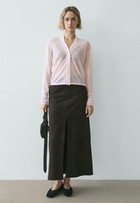 Cardigan rose clair transparent avec boutons, associé à une jupe en daim marron avec une fente à l'avant. Des ballerines noires et un petit sac complètent la tenue.