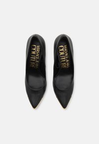 Scarpe con tacco alto in pelle nera con punta affusolata, caratterizzate da una superficie liscia e logo "VERSACE JEANS COUTURE" stampato in oro all'interno.