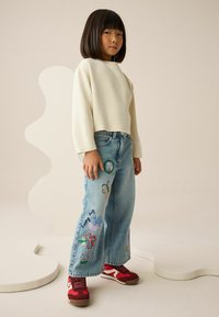 Kremowa krótka bluza z długim rękawem, jasnoniebieskie jeansy z kolorową haftą oraz czerwone trampki z ciemnymi dodatkami.