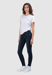 Wit katoenen T-shirt met korte mouwen en een rood logo op de borst, gecombineerd met donkerblauwe skinny jeans en witte sneakers.