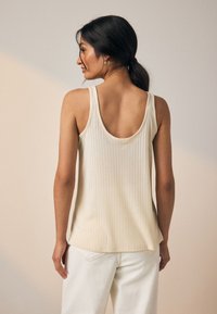 Débardeur côtelé crème avec un dos échancré, doté de larges bretelles et d'une texture douce et subtile. Porté avec un jean de couleur claire.