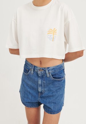 Person trägt ein weißes, bauchfreies T-Shirt mit gelbem Palmen- und blauem Wellenmotiv, kombiniert mit hoch taillierten blauen Jeansshorts.