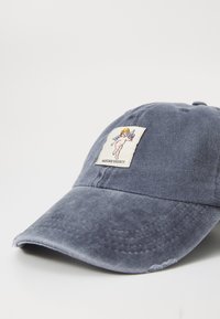 Denim baseball cap in faded navy with a curved brim. Features a white label patch depicting an illustration. Distressed edge detailing.
