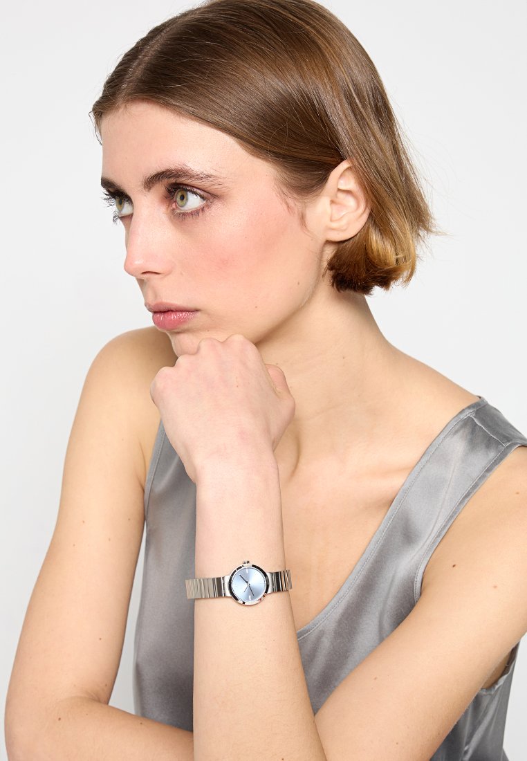 Zilveren metalen horloge met een ronde blauwe wijzerplaat, met een minimalistisch ontwerp en een mesh band, gedragen om een pols.