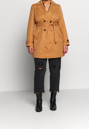 Personne portant un trench-coat beige ceinturé, un jean noir court et déchiré, ainsi que des bottines noires, se tenant devant un fond clair uni.