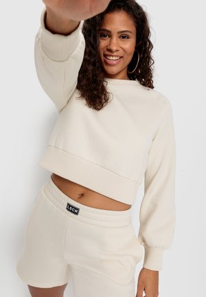 Felpa beige a maniche lunghe con polsini a coste, abbinata a pantaloni shorts in tinta a vita alta. Tessuto morbido con vestibilità rilassata e dettaglio con logo sui pantaloni.