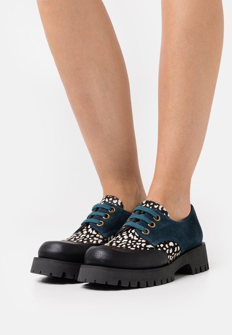 Chaussures oxford à semelles épaisses noires avec panneaux en daim bleu sarcelle et tissu à pois noir et blanc, portées sur des jambes nues.