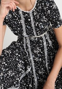 Svart blommig klänning med små vita och blå blommor, spetsdetalj längs halsringningen och vertikal panelering, med midja som framhäver figuren och en svängd kjol.