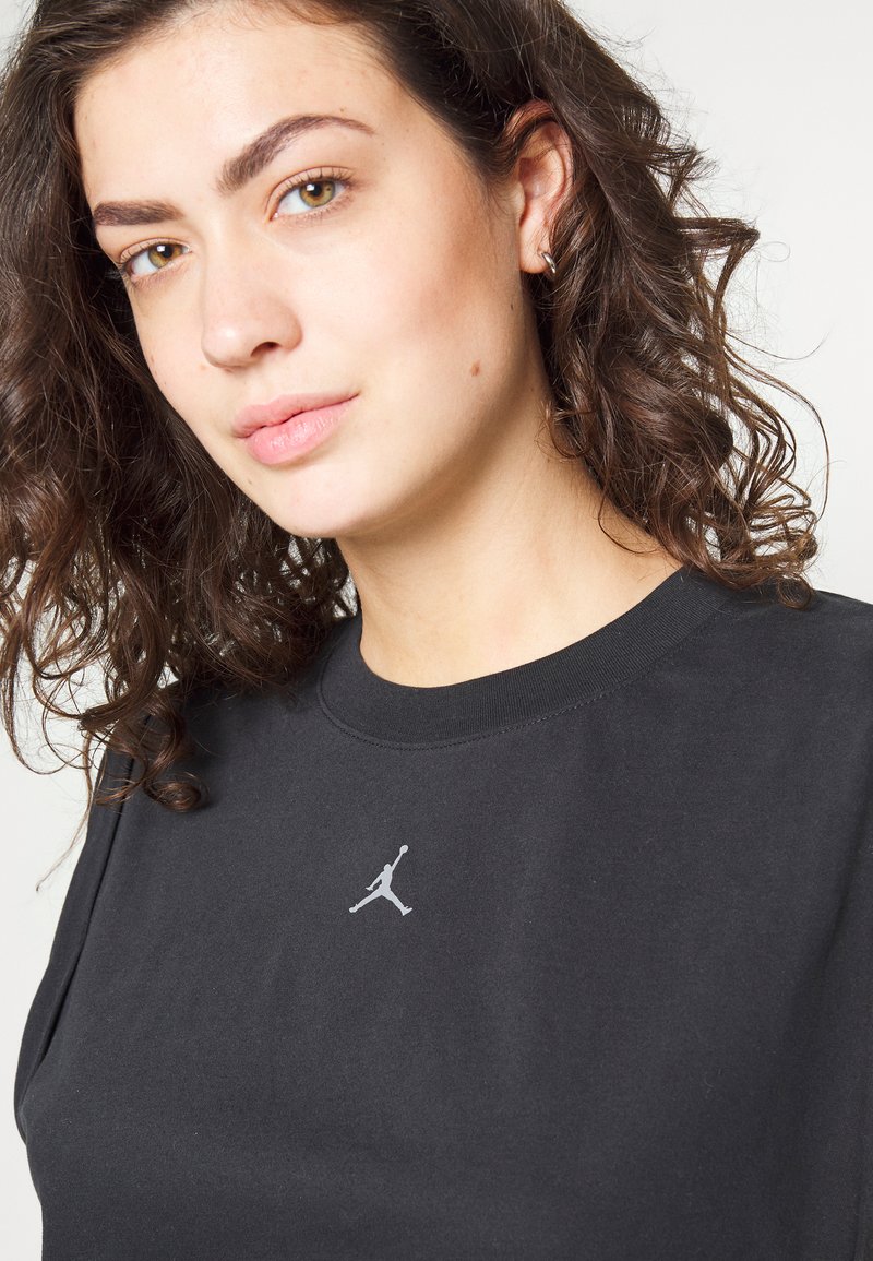 Młoda kobieta z kręconymi brązowymi włosami, ubrana w czarną koszulę z małym białym logo Jumpman na piersi, na jednolitym tle.