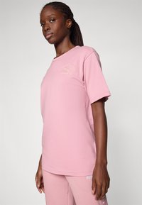 Hellrosa T-Shirt mit kurzen Ärmeln, aus weichem Baumwollstoff, mit rundem Halsauschnitt und goldenem gesticktem Logo. Glatte Textur, lässige Passform.