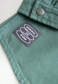 Material verde cu finisaj texturat, cu o etichetă dreptunghiulară neagră cu text alb „MAKE IT FUN” și un accent metalic sub formă de nasture.