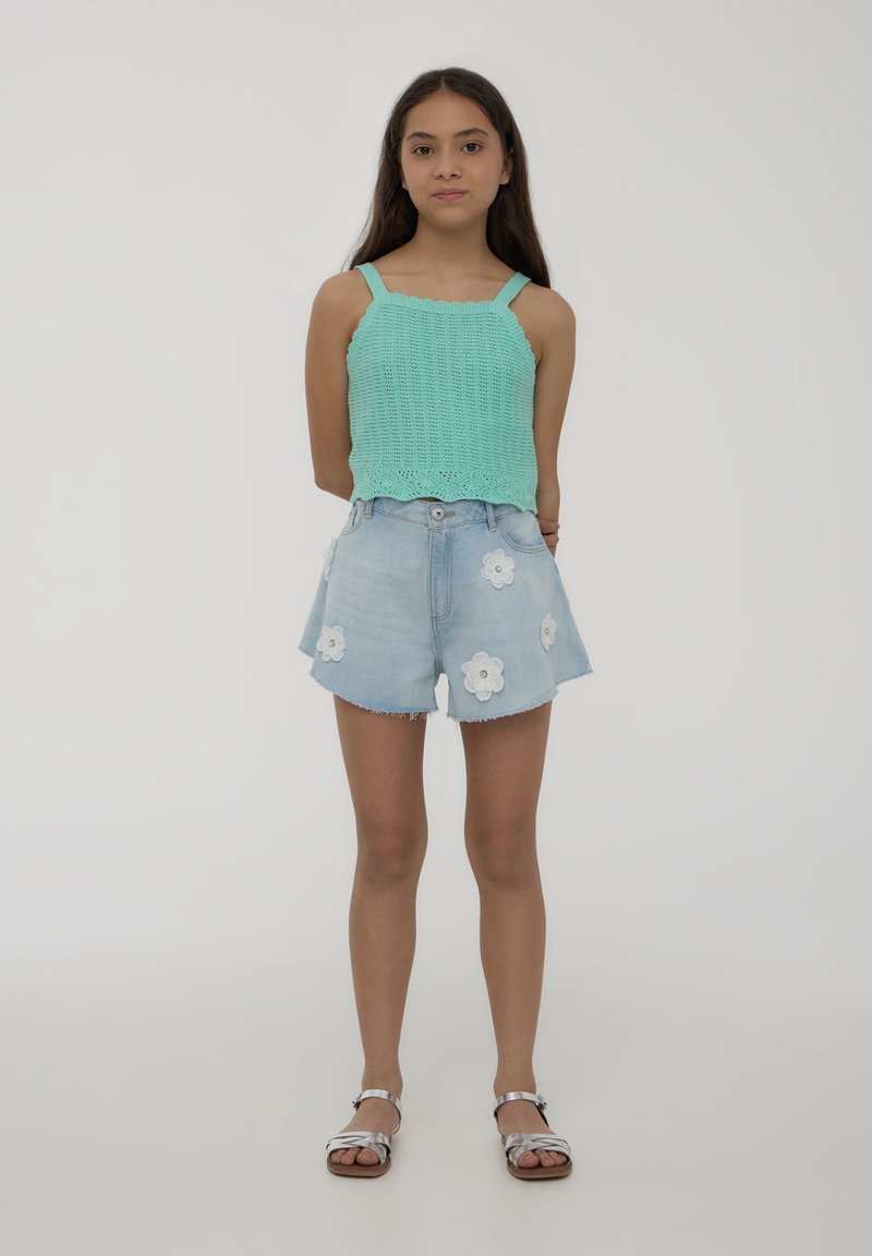 Top in maglia verde menta con bordi scallop, abbinato a pantaloni shorts in denim azzurro chiaro con applicazioni floreali bianche. Sandali argentati completano il look.