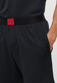 Shorts noirs avec une texture lisse, dotés d'une ceinture élastique noire et d'une étiquette rouge bien visible portant le mot "HUGO".