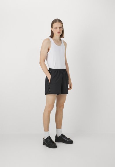Black shorts with elastic waistband and side pockets, paired with a fitted white tank top. Model wears black sneakers and white socks.