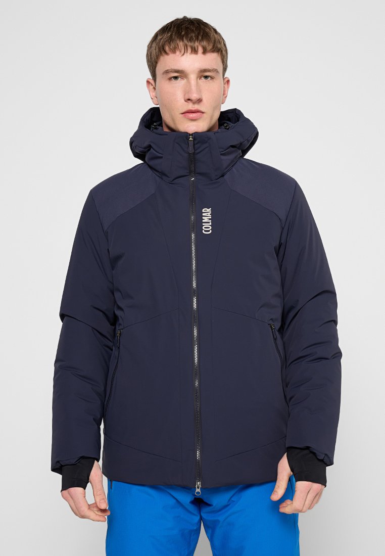 Giacca isolante blu navy con cappuccio, dotata di zip frontale, tasche laterali e dettaglio logo sul petto. Texture liscia, design sportivo.