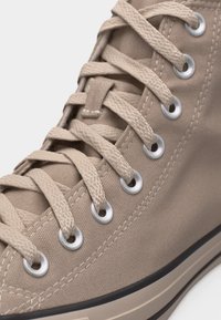 Κοντινό πλάνο σε μπεζ πάνινο αθλητικό παπούτσι υψηλής κοπής με μπεζ κορδόνια περασμένα από ασημί μεταλλικές τρύπες και λευκή λαστιχένια σόλα με μαύρη ρίγα.