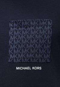 Μπλουζάκι σε ναυτικό μπλε με ραμμένο σχέδιο πατς που επαναλαμβάνει το λογότυπο «MK» και το κείμενο «MICHAEL KORS» σε λευκό στο κάτω μέρος. Μαλακό βαμβακερό υλικό.