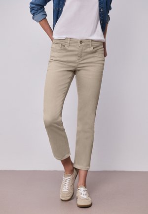 Femme portant un pantalon beige slim court, des baskets blanches, un t-shirt blanc et une veste en jean avec les manches retroussées, debout devant un fond uni.