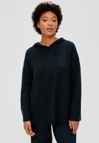 Dunkelgrüner, übergroßer Hoodie aus weichem Stoff, mit einer Kapuze mit Kordelzug, gerippten Bündchen und Saum. Das Model trägt dunkelblaue Hosen.