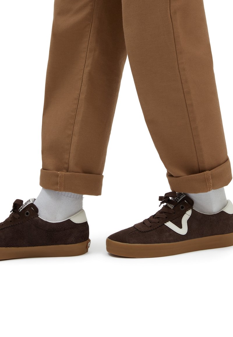 Jambes portant un pantalon marron retroussé, des chaussettes blanches et des baskets marron foncé avec des semelles en gomme et des accents blancs.