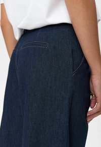 Back view of person wearing dark blue denim pants with a back welt pocket and a white tucked-in top.