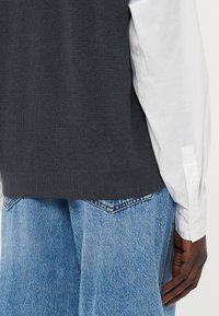 Grå strikket vest lagt over en hvit skjorte, parret med lyseblå denim. Vesten har en teksturert finish og ribbet kant.