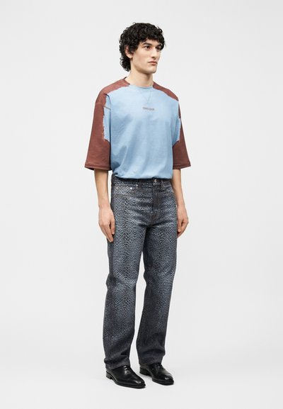 Νεαρός άνδρας που στέκεται, φορώντας μια φαρδιά ανοιχτό μπλε και καφέ κοντομάνικη πουκάμισα, παντελόνι με γκρι σχέδιο και μαύρα επίσημα παπούτσια, με απλό φόντο.