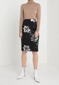 Beige pletený rolák doplněný černou tužkovou sukní s květinovým vzorem, která má bílé lilie. Modelka má na sobě bílé kotníkové boty s černými pruhy.