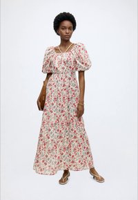 Femme portant une longue robe blanche à fleurs avec des manches bouffantes, des sandales tongs, et portant un sac à main marron sur un fond uni.