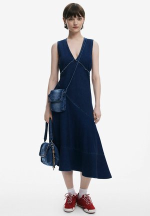 Abito in denim con profondo scollo a V, design avvolgente e orlo svasato. Abbinato a una borsa a tracolla in denim di piccole dimensioni e sneakers rosse.