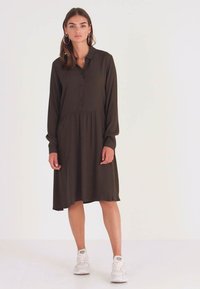 Vestido de manga longa, com botões, em tecido verde escuro; apresenta um corpete justo com uma saia rodada e uma bainha sem costura. Usado com ténis brancos.