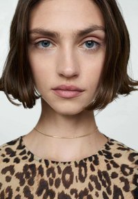 Model cu părul scurt, brunet, purtând un tricou cu imprimeu de leopard. Are ochi albaștri și o brățară subțire din aur. Ten neted; culoare neutră a buzelor.