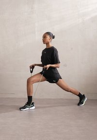 Black athletic shirt and shorts, black sneakers with green accents, and resistance bands. Model in a lunge position on a smooth surface.
