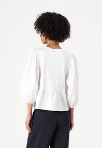 White blouse with puff sleeves, gathered hem, and a rounded neckline. Soft fabric with subtle sheen, featuring a small logo on the back.