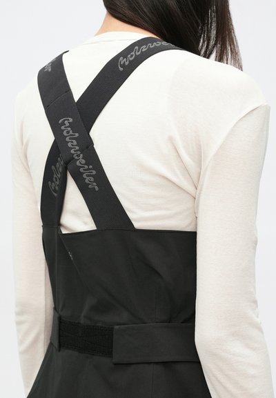 Schwarze Overalls mit dicken, überkreuzten Trägern und "Holzweiler"-Markenlogo, kombiniert mit einem hellbeigen Langarmshirt. Dezente Texturen im Stoff.