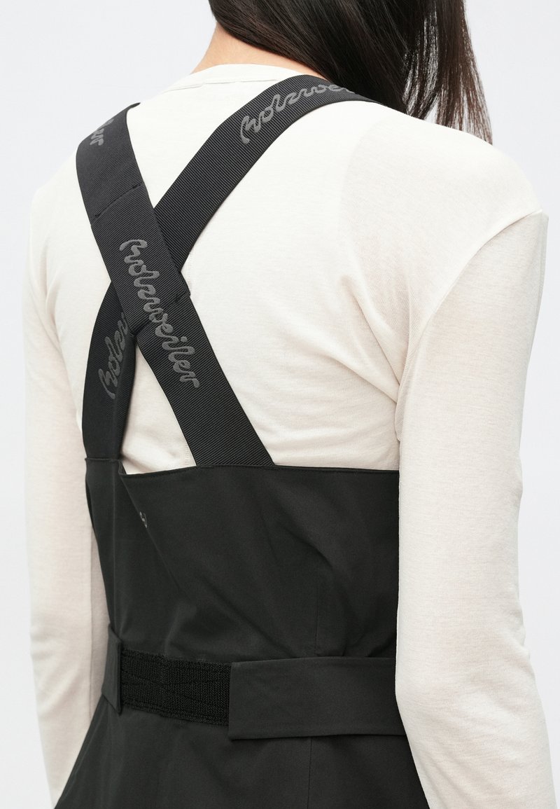 Black overalls with thick, crossback straps featuring "Holzweiler" branding, paired with a light beige long-sleeve shirt. Subtle textures in fabric.