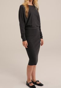 Femme portant une robe longue ajustée gris foncé avec une jupe crayon midi et des chaussures Mary Jane noires, se tenant devant un fond uni.