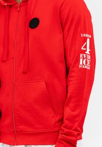 Rote Kapuzenjacke mit Reißverschluss und rundem schwarzem Logo auf der Brust. Verfügt über weißen Text auf dem linken Ärmel, Kängurutaschen und verstellbare Zugbänder.