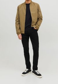 Khakifarbene Bomberjacke mit einer Reißverschlusstasche, navyfarbener Pullover darunter, schwarze Slim-Fit-Jeans und schwarze Sneakers mit weißen Schnürsenkeln.