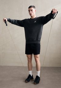 Black sweatshirt with logo, black shorts, black sneakers with gum soles, holding resistance bands, standing on a neutral floor against a light wall.