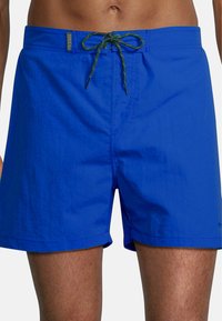 Shorts de bain bleu vif avec une finition texturée, présentant une couleur unie, une taille élastique avec un cordon de serrage vert et une étiquette de logo.