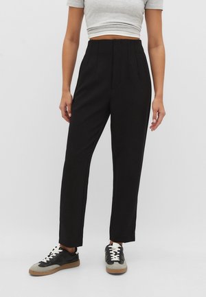 Mujer que lleva pantalones negros de cintura alta y pierna recta, camiseta gris corta y zapatillas negras y grises con cordones blancos, de pie contra un fondo liso.